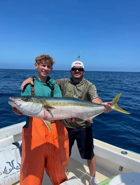 Two anglers holding a bright yellowtail over calm blue offshore water.